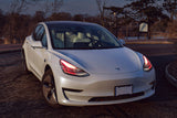 Three quarters view of a Tesla Model 3 with red headlight DRLs.